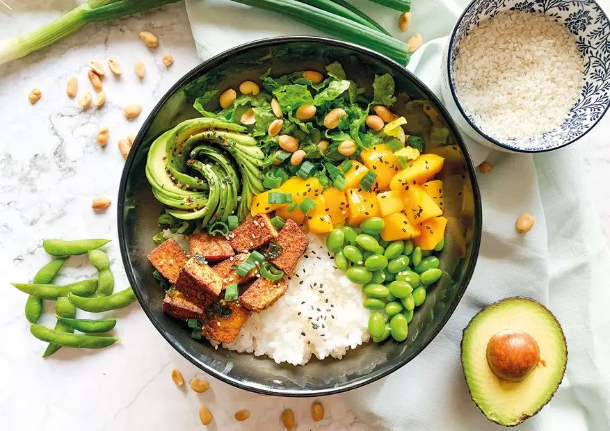 A balanced plate shown from above — a bowl of rice, seasoned tofu, mango, edamame, avocado, peanuts, and fresh greens.